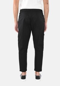 Pantaloni da jogger neri con cintura elastica, gambe tapered e tasche laterali, realizzati in tessuto liscio. Indossati con scarpe nere senza lacci.