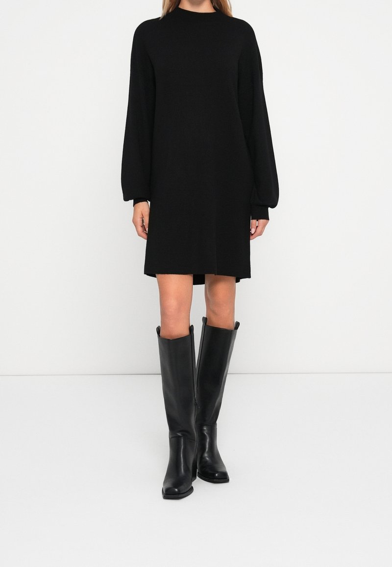 Abito nero in maglia con colletto alto e maniche lunghe e voluminose, abbinato a stivali neri in pelle al ginocchio con un design elegante e aderente.