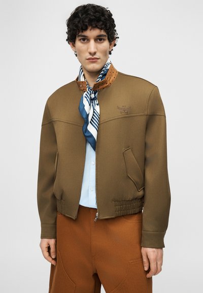 Άνδρας με σγουρά μαλλιά, που φορά καφέ μπουφάν τύπου μπόμερ, ανοιχτό γαλάζιο πουκάμισο, ριγέ μπλε και λευκό κασκόλ και σκουρόχρωμο παντελόνι στοιχισμένος μπροστά σε απλό φόντο.