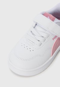 Zapatilla deportiva blanca de cuero con acentos rosas y una correa de velcro. Presenta puntera redonda, detalle de cordones y pequeños agujeros de ventilación en la puntera.