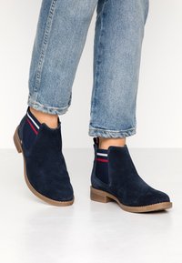 Botines de ante azul marino con paneles laterales elásticos que presentan rayas rojas y blancas; suela marrón claro; punta redondeada; diseño casual.