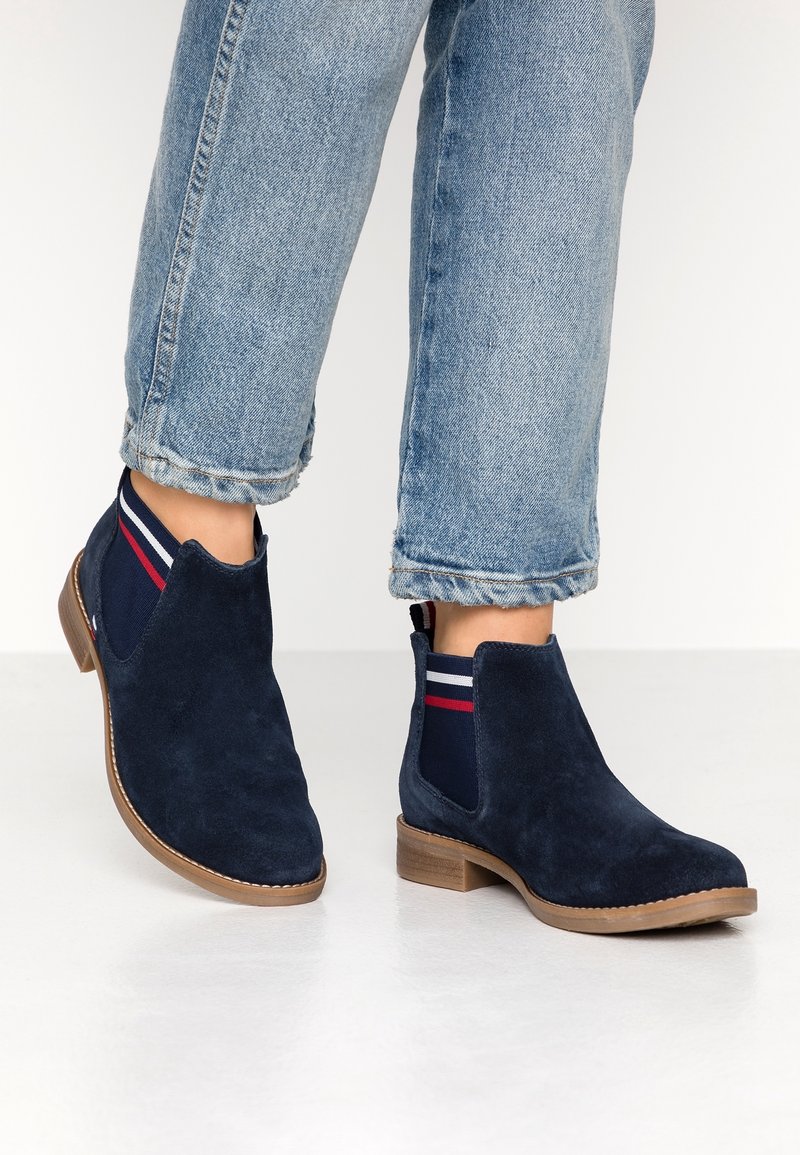 Botines de ante azul marino con paneles laterales elásticos que presentan rayas rojas y blancas; suela marrón claro; punta redondeada; diseño casual.