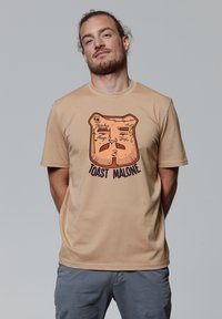 Beige katoenen t-shirt met een afbeelding van een geroosterde sneetje brood met gezichtskenmerken en de tekst "TOAST MALONE" in donkere letters.