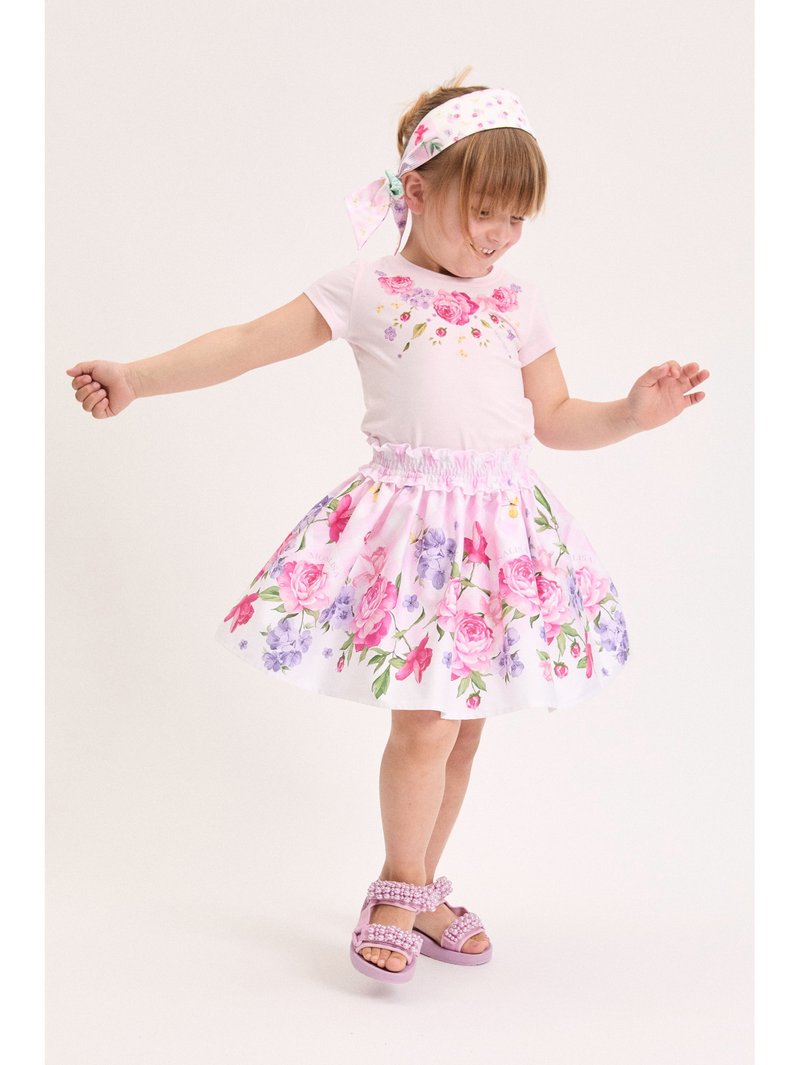 Νεαρή κοπέλα που χορεύει φορώντας ένα ροζ φόρεμα με λουλούδια, ταίριαστο κορδέλα και ροζ σανδάλια με μαργαριτάρια, σε απλό φόντο.