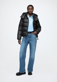 Czarna kurtka puchowa z kapturem, noszona na jasnoniebieskiej koszulce polo. Jasnoniebieskie dżinsy i czarne buty na masywnym spodzie dopełniają strój.