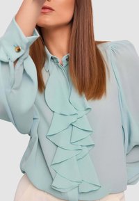Blusa azzurro chiaro in tessuto liscio, con volant sul davanti, maniche lunghe con dettagli a bottoni e scollo con colletto.