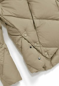 Beige gewatteerde jas met diagonale stiksels, drukknoopsluitingen en een gedeeltelijk geopende rits op een witte achtergrond.