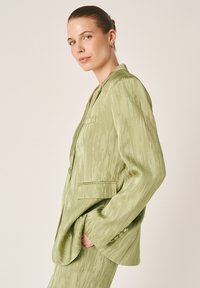 Frau in hellgrünem, strukturiertem Blazer mit Taschen, im Profil stehend mit Hand in der Tasche, neutraler Hintergrund.