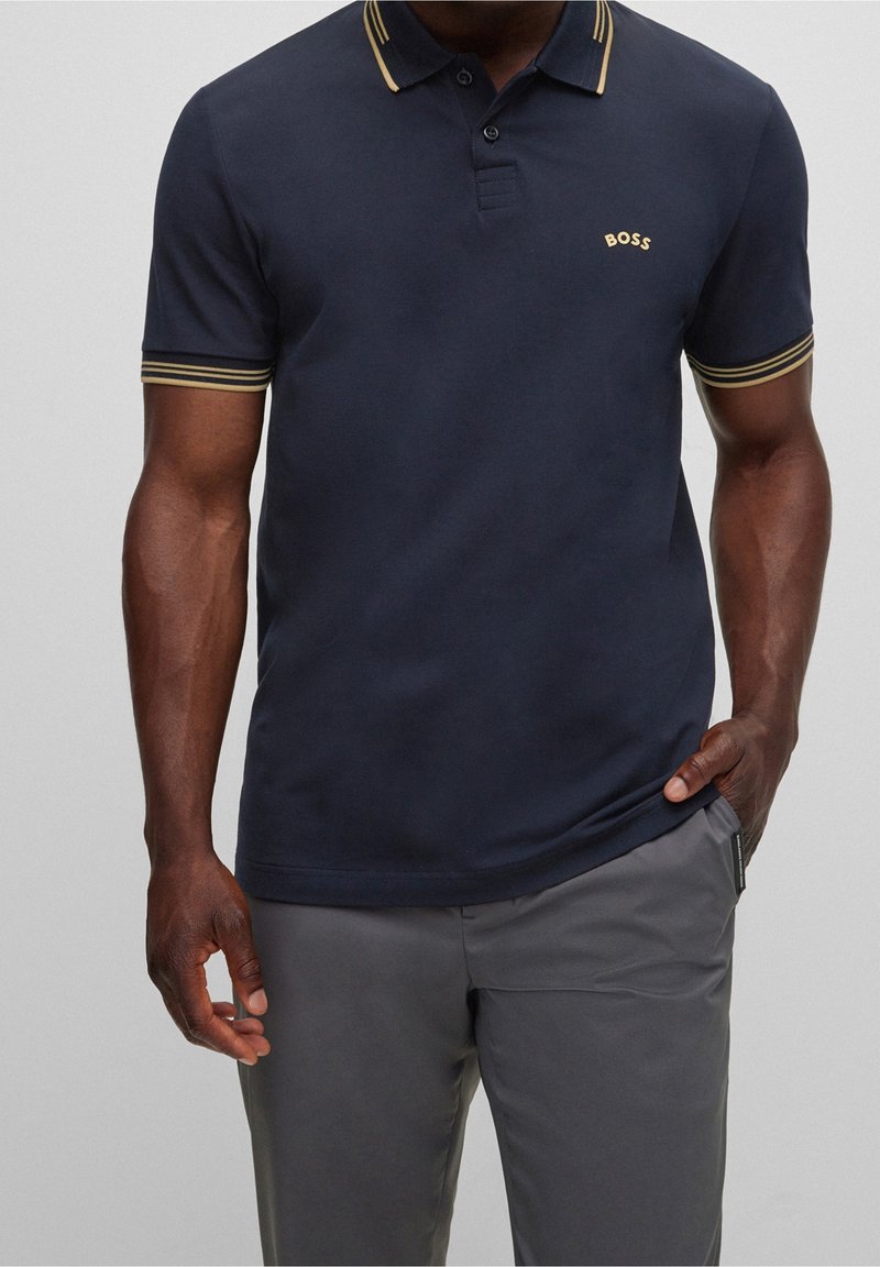 Marineblaues Poloshirt mit Zwei-Knopf-Leiste, gold gestreiftem Kragen und Ärmelsäumen, mit eingesticktem "BOSS"-Logo. Kombiniert mit grauer Hose.