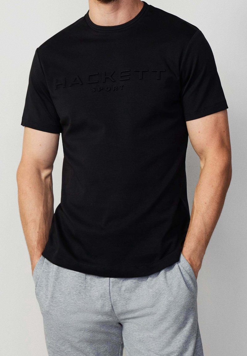 Czarna koszulka polo z krótkim rękawem z wypukłym logo "HACKETT SPORT" z przodu, połączona z szarymi spodniami ze sznurkiem. Gładka faktura materiału.