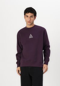 Violetinis džemperis su apvalia kaklo linija, raukšlėmis ant rankovių ir apačios, su maža balta trikampio forma logo ant krūtinės.