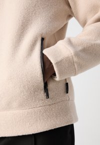 Beige fleece pullover with side zip pockets, ribbed cuffs, and a straight hem. Tags visible on the side. Soft texture, relaxed fit.