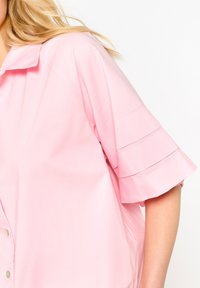 Pink short-sleeve shirt made of lightweight fabric. Features a button front, collar, and pleated detailing on sleeves for added texture.