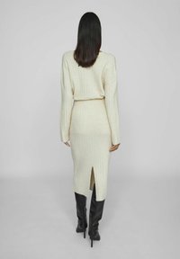 Vestido de punto acanalado crema con cintura ceñida, mangas largas y una abertura en la espalda. Combinado con botas de tacón alto negras, exhibiendo una textura suave.