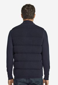 Mörkblå stickad väst med horisontell ribbad textur, ståkrage och vadderade sektioner på baksidan. Bärs över en långärmad skjorta.