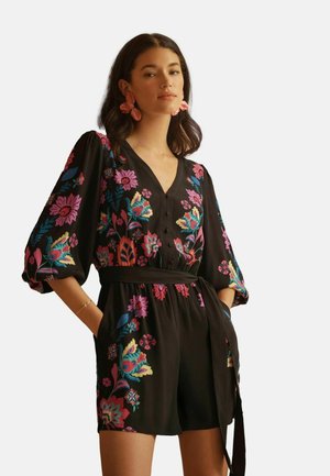 Frau trägt einen schwarzen, geblümten Jumpsuit mit Puffärmeln, V-Ausschnitt und einem gebundenen Taillengürtel, accessoriert mit großen pinken Blumenohrringen.