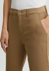 Gros plan sur une personne portant un pantalon marron avec un bouton et une fermeture éclair à l'avant, une main dans la poche, et un haut blanc rentré.