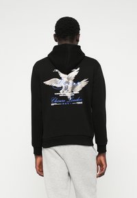 Svart hettegenser laget av bomullsblanding, med en stor hvit fuglegrafikk og blå tekst på baksiden, med snor i hetten og ribbede mansjetter.