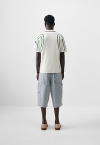 Light cream short-sleeve sweater with green outline detailing, paired with loose denim shorts. Model wears textured tan slip-on shoes.