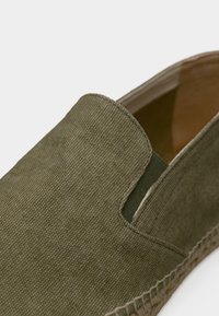 Πράσινο-ελιάς πανί slip-on παπούτσι με ελαστικό πλευρικό πάνελ και πλεγμένη σόλα από τζούτ, παρουσιάζεται από κοντινή γωνία.