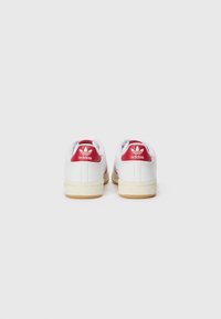 Zapatillas Adidas blancas con acentos rojos, con un exterior de cuero suave, tres rayas rojas y una suela de goma. Logo en el talón.