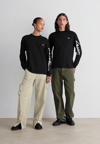 Schwarze Langarmshirts mit grafischem Text auf den Ärmeln, kombiniert mit beigen Cargo-Hosen und dunkelgrünen Hosen, beide getragen mit schwarzen Sneakers.