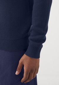 Marineblå strikket sweater med ribbede manchetter og kant, der har et tekstureret mønster. Hånd hviler på et mørkeblåt beklædningsgenstand.