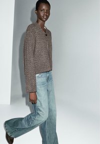 Bruine, geribbelde gebreide cardigan met een V-hals en knoopsluiting, gecombineerd met lichtblauwe, wijdvallende jeans. Model staat op een effen achtergrond.