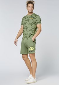 Grünes Baumwoll-T-Shirt mit schwarzem Tierprint und gelbem Text; kombiniert mit grünen Shorts mit einem gelben Sternlogo und schwarzem Text. Weiße Sneakers.