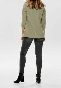 Olive green tailored blazer with three-quarter sleeves, paired with dark grey skinny jeans and black ankle boots. Smooth texture, sleek design.