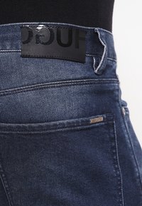 Vaqueros de mezclilla azul oscuro con un parche de cuero negro en la cintura, costuras en contraste y una pequeña etiqueta metálica en el bolsillo.