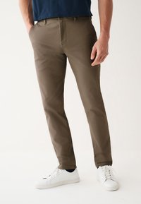 Brown, slim-fit trousers made of smooth fabric with front pockets, styled with a zipper fly, paired with white sneakers.