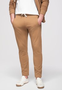 Personne portant un pantalon de jogging beige, des baskets blanches, une chemise blanche et une veste zippée beige, debout les mains dans les poches sur un fond uni.