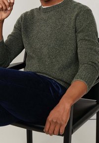 Personne assise sur une chaise noire portant un pull gris en maille et un pantalon en velours côtelé bleu foncé, avec un bras reposant sur l'accoudoir.