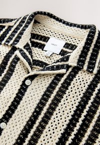 Camisa de tejido con botones, base color crema con rayas negras, tela texturizada, cuello y una etiqueta en el centro con la marca "NEXT".