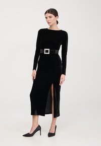 Abito lungo in velluto nero con maniche lunghe e spacco laterale, vita cinchata con una fibbia quadrata decorativa, abbinato a tacchi alti neri scintillanti.