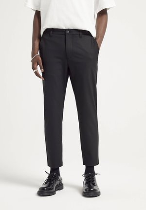 Homme portant un pantalon noir slim avec passants de ceinture, des chaussettes noires, des chaussures noires à lacets, un haut blanc à manches courtes, et un bracelet et une bague argentés.