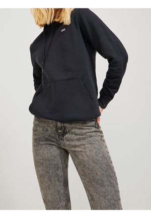 Vrouw draagt een zwarte hoodie met het logo "JJXX" en een grijze acid-wash jeans met hoge taille, staand tegen een effen lichte achtergrond.