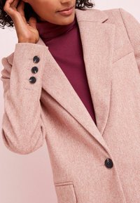 Růžový blejzr s texturovanou látkou, který má klopový límec, tři černé knoflíky na rukávu a přední kapsu.