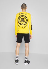 Camisa amarilla de manga larga con diseños gráficos negros en la parte posterior y en las mangas, combinada con pantalones cortos negros y zapatillas deportivas.
