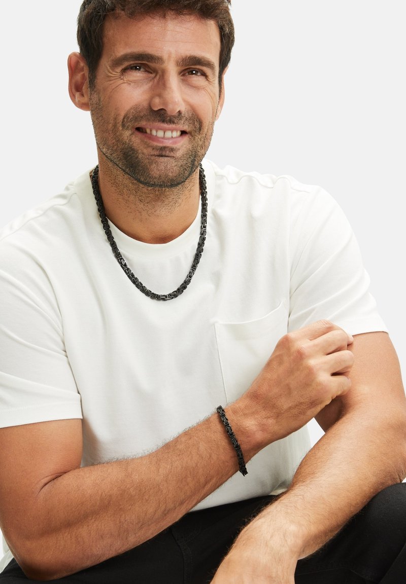 Zwarte metalen ketting en armband gedragen door een man, met een witte t-shirt met korte mouwen en een borstzak.