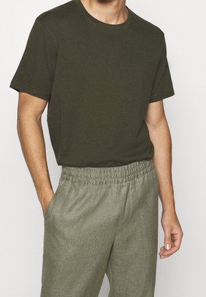 Mand iført en olivengrøn t-shirt med korte ærmer, stukket ned i lysegrå elastiske bukser med hænderne afslappet ned langs siden.