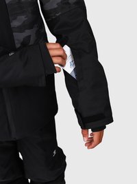 Eine Hand schiebt einen Skipass in eine schwarze Taschenhülle mit einem Klettverschluss an einer Jacke mit schwarzem Muster.