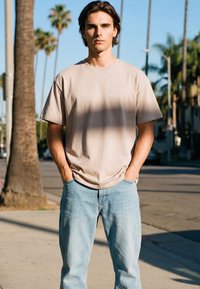 Mladenič s srednje dolgimi rjavimi lasmi stoji z rokami v žepih, oblečen v bež majico in svetlo modre kavbojke na sončni ulici s palmami.