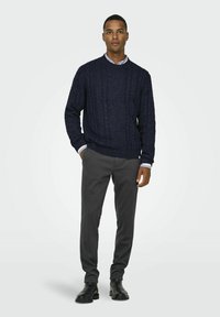Marineblauwe kabelgebreide trui met geribbelde manchetten en zoom, gecombineerd met grijze slim-fit broek. Het model draagt zwarte enkellaarsjes.