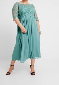 Tealfärgad klänning i knälängd med spetsiga trekvartslånga ärmar och samlad midja, tillverkad av lättviktsmaterial. Har en svävande kjol och svarta klackskor.