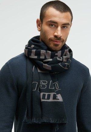 Black scarf with gray lettering, made of soft fabric, featuring a fringe edge. Worn around the neck over a textured navy sweater.