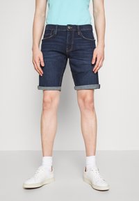 Shorts en denim bleu foncé avec des ourlets retournés, dotés de poches avant et d'une fermeture à boutons. Assortis à des baskets blanches et des chaussettes de longueur mi-mollet.