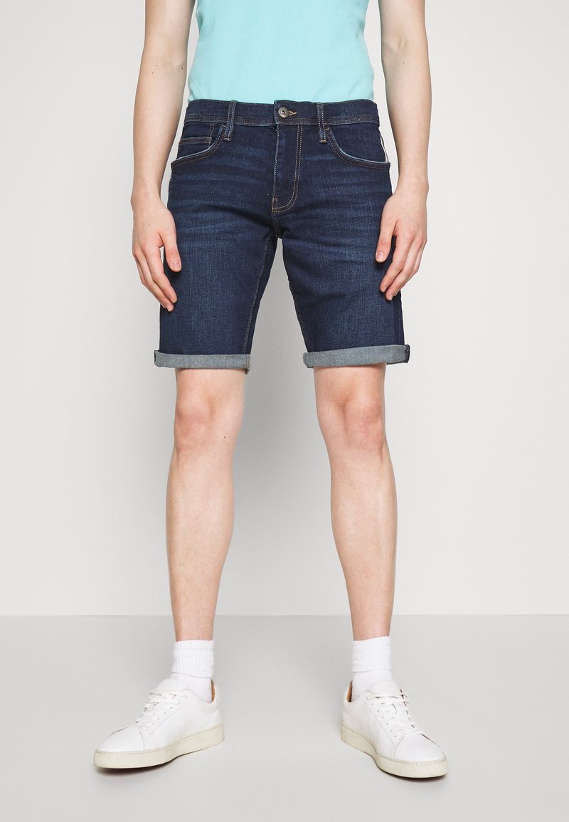 Shorts en denim bleu foncé avec des ourlets retournés, dotés de poches avant et d'une fermeture à boutons. Assortis à des baskets blanches et des chaussettes de longueur mi-mollet.