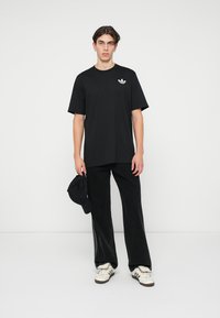 Νεαρός άνδρας που στέκεται στον τοίχο λευκού χρώματος, φορώντας μαύρο μπλουζάκι της Adidas, μαύρο παντελόνι, λευκά sneakers της Adidas, κρατώντας ένα μαύρο καπέλο στο αριστερό χέρι.